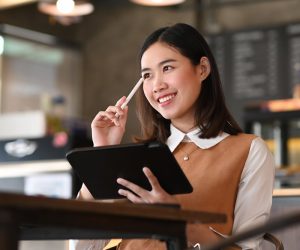 Smiling young female designer holding digital tablet and thinking about her project while sitting in modern office.